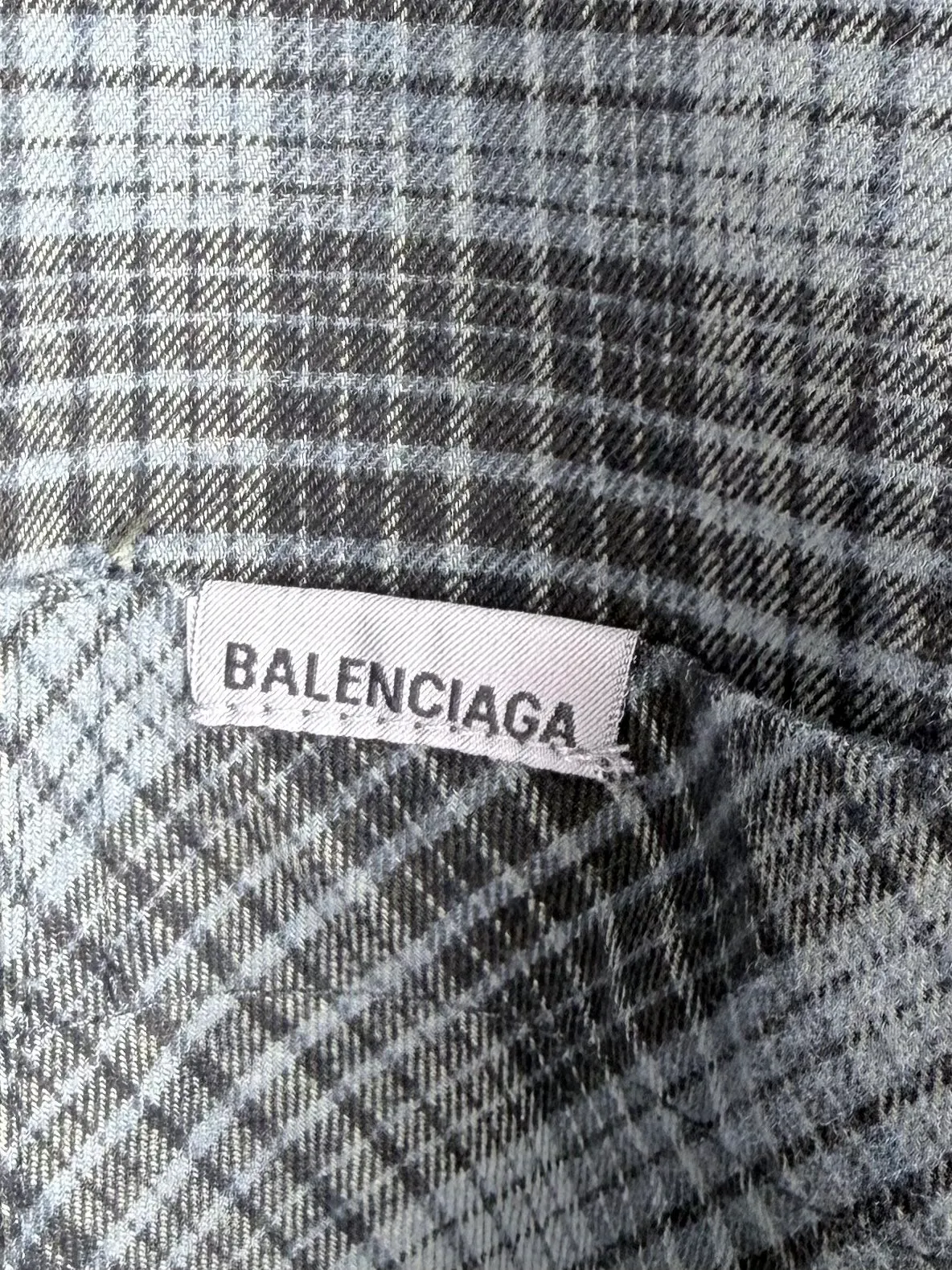 BALENCIAGA/balenciaga blue and gray plaid zipper pocket logo quilted jacket combines a three-layer structure of plaid woven fabric, filling cotton, and lining, and integrates plaid alignment technology and quilted garment production standards. There are multiple process difficulties in the whole process. The surface layer of the body is made of blue-gray Retro plaid fabric, and the cutting machine production strictly follows the global grid standards. The arrangement direction of the cutting pieces is fixed and cannot be changed at will. The horizontal and vertical lines of shoulder seams, side seams, and zipper placket joints must be consistent and coherent. The left chest patch pocket is cut independently, and the bag check pattern must be aligned with the front reference pattern. Slight deviations will destroy the visual integrity and directly increase fabric loss and cutting time. The most difficult part of the style is the three-layer composite sewing. The outer plaid fabric, filling cotton, and lining fabric have different physical properties, and the preshrinking processing parameters need to be adjusted separately. Pressure and temperature control are crucial during lamination operations. Once the parameters are out of balance, it can easily cause fabric stretch and distortion, plaid dislocation, and wrinkles and bulges in garments. At the same time, it is necessary to reasonably plan the quilting line to avoid the visual beauty of the grid pattern from being cut by the lines on the basis of firmly fixing the cotton layer and preventing cotton from running away. The placket adopts a centered zipper structure. It is difficult to sew the zipper tape and the quilted panels. It is required that the width of the left and right plackets be equal, the zipper routing is straight and smooth, and there is no lag in opening and closing. When the shirt lapel is filled with cotton, the thickness of the cotton needs to be controlled to prevent the collar from becoming bloated and unnaturally folded. The metal nameplate of the chest pocket is sewn at fixed points to avoid pulling the fabric during the operation and ensure that the plaid pattern is smooth and not deformed. The quilted material increases the overall thickness of the fabric. When sewing, you need to continuously adjust the presser foot pressure and stitch length to adapt to changes in sandwich thickness. The low-temperature and gentle ironing process is used for post-finishing to prevent the plaid from fading and the cotton layer from clumping due to high temperatures. From the precise alignment of cutting pieces, three-layer fabric pretreatment, composite quilting to garment finalization inspection, the processes are interlocking and the error tolerance rate is extremely low, testing the accuracy of plate making, equipment control and turning technology. Size:XS S M L